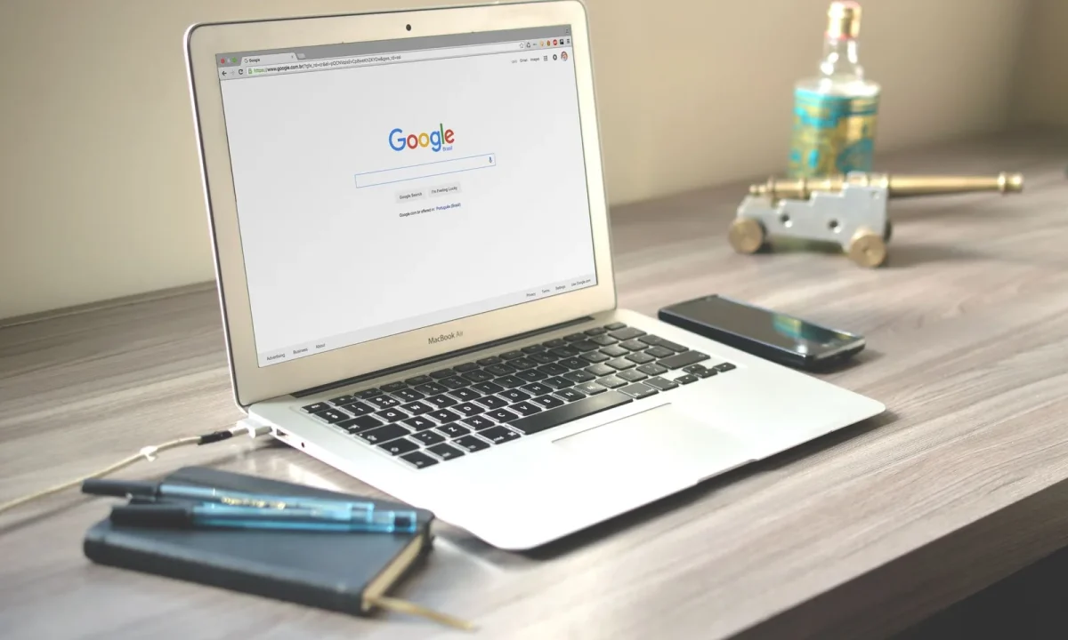 Espaço de trabalho organizado com laptop mostrando busca no Google, smartphone e caderno sobre mesa de madeira