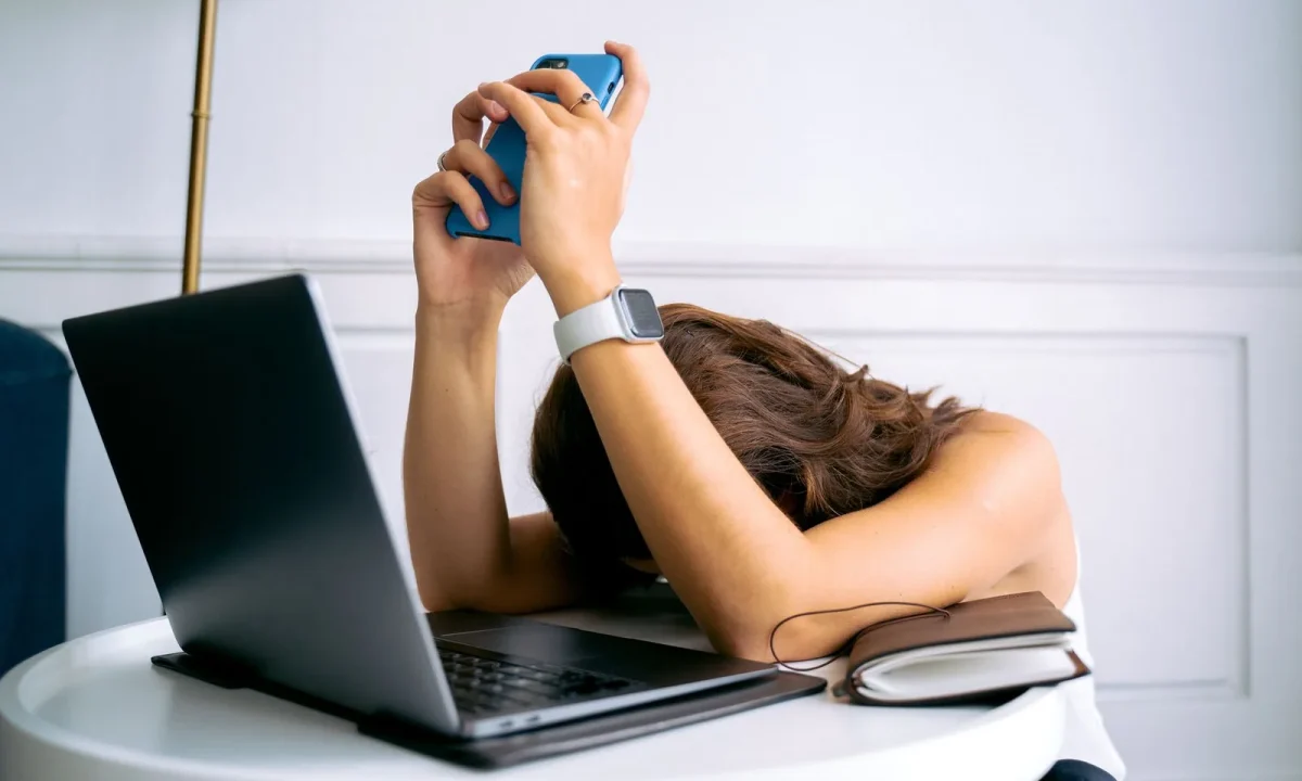 Mulher sobrecarregada com trabalho, descansando a cabeça na mesa com laptop, celular e smartwatch, simbolizando falta de