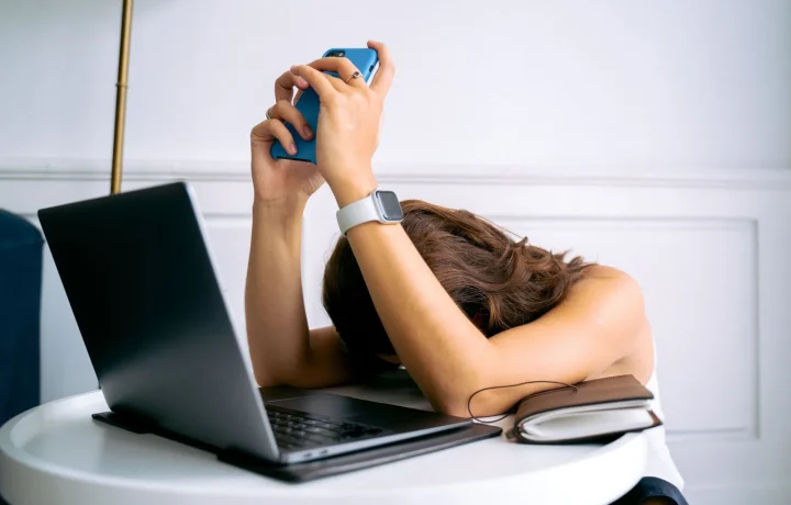 Mulher sobrecarregada com trabalho, descansando a cabeça na mesa com laptop, celular e smartwatch, simbolizando falta de