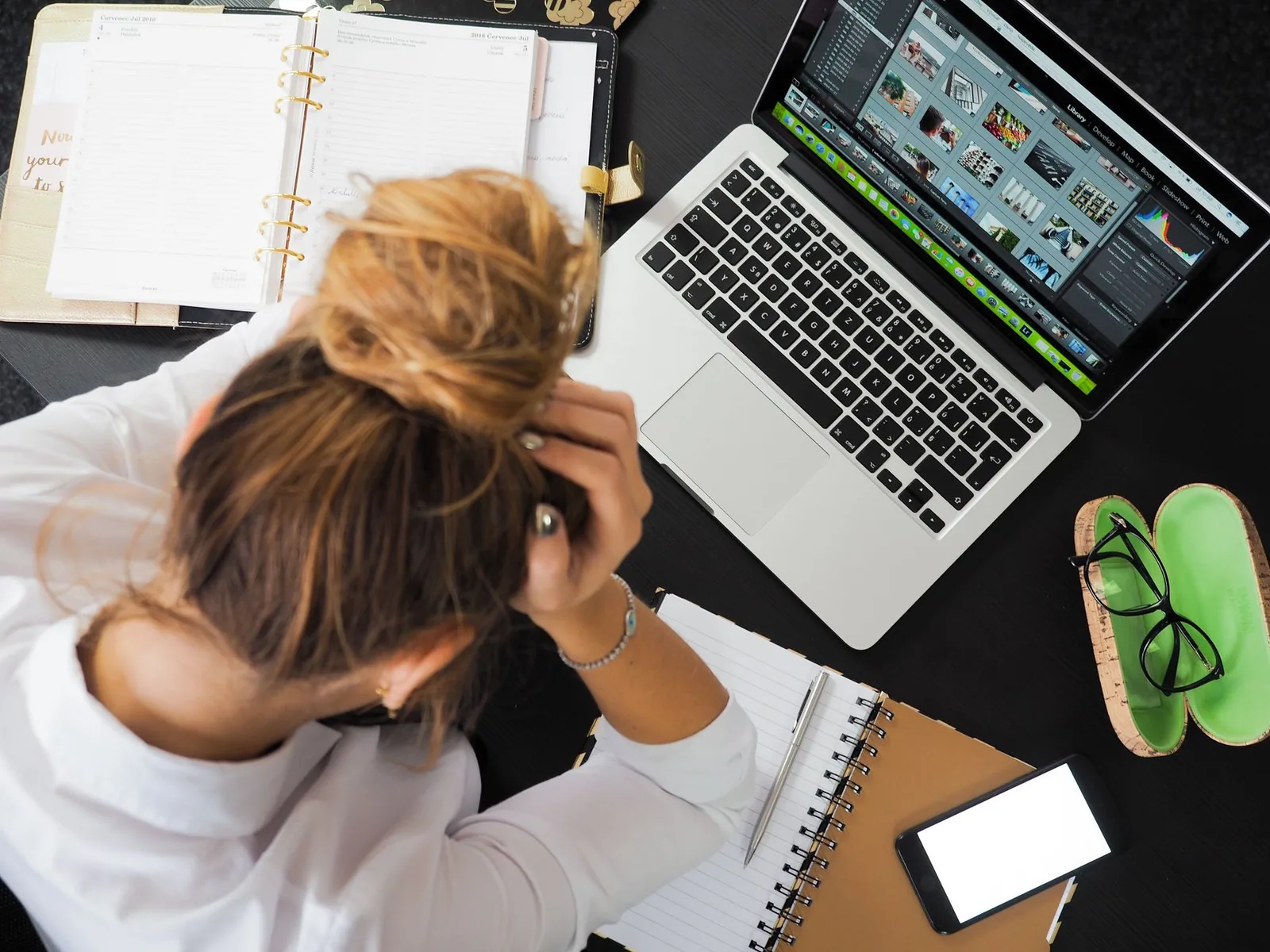 Vista aérea de uma mulher estressada trabalhando em uma mesa com laptop, celular e cadernos, ilustrando auditoria de site